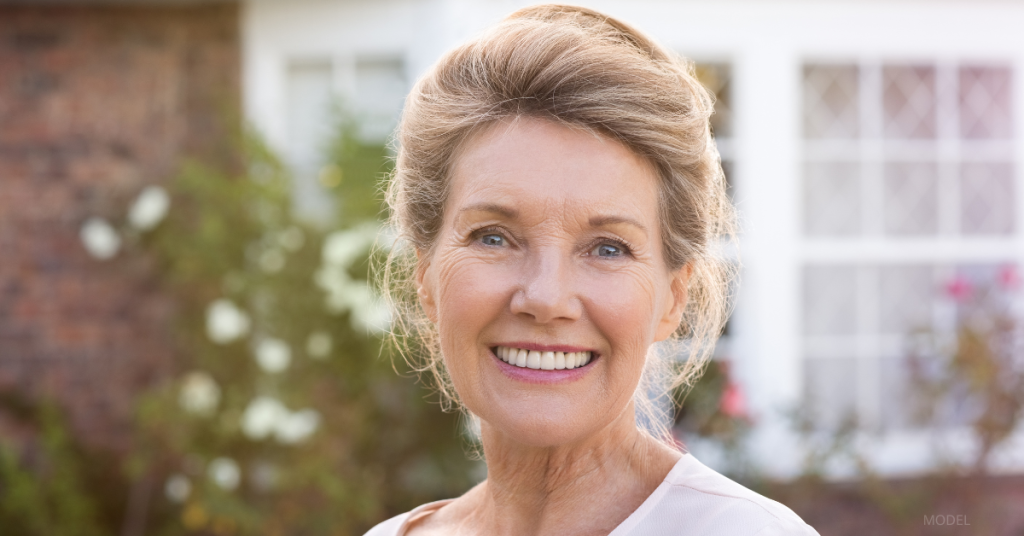 An older woman with light colored hair pulled back is smiling and looking forward. (model)